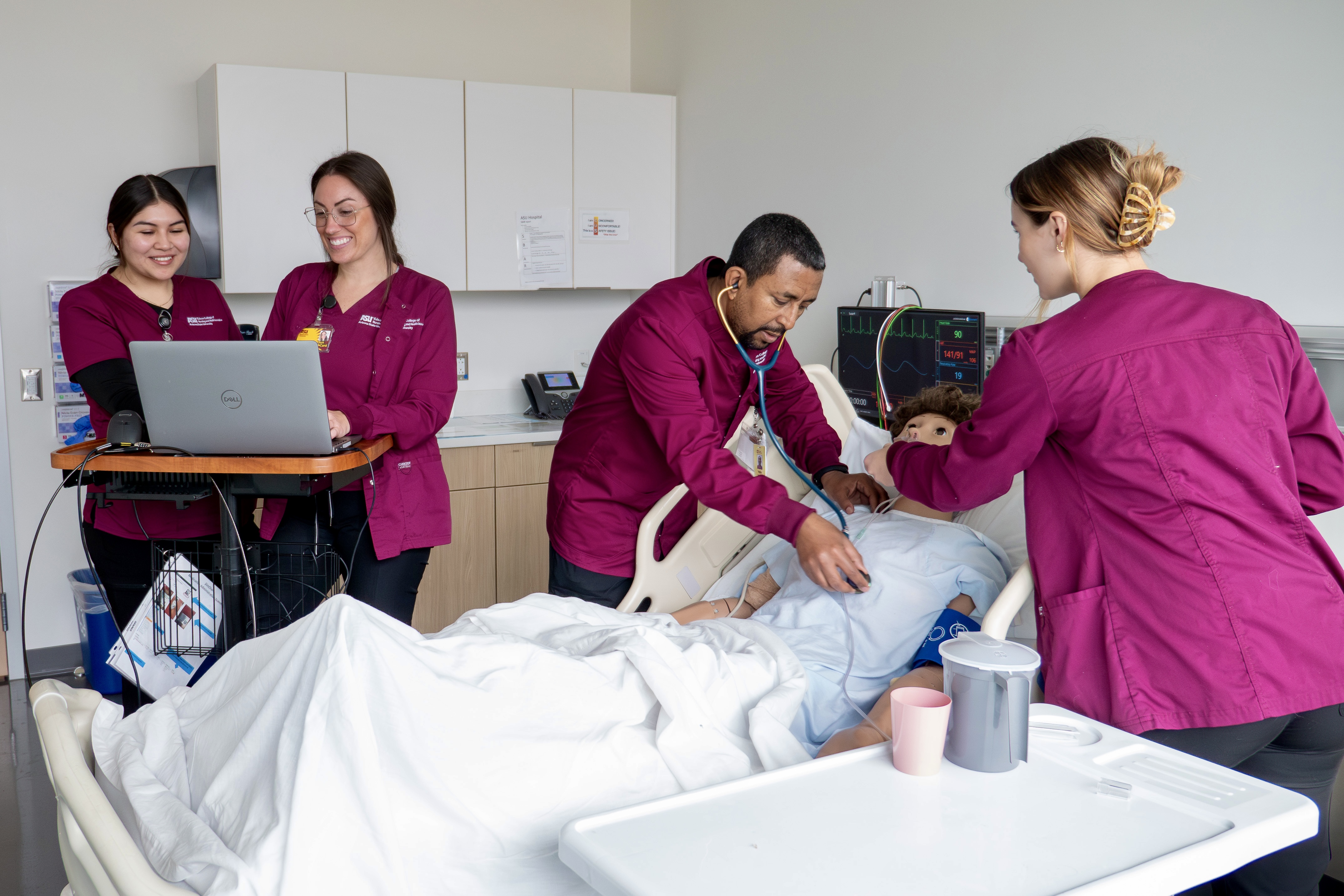 Four Edson College nursing students learning and interacting with a medical training mannequin in a clinical simulation setting.